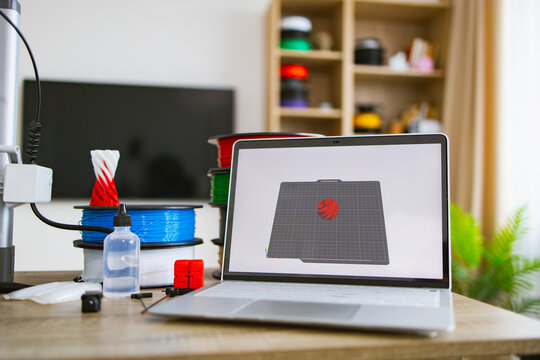 A laptop displays a 3D model next to spools of filament, a 3D printer, and accessories, ready for creation