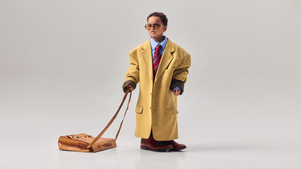 Little boy wearing oversized coat and sunglasses dragging large leather briefcase across studio floor. Concept of childhood career dreams, business ambition, leadership imagination, playful role play. © Lustre Art Group 