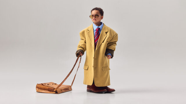 Little boy wearing oversized coat and sunglasses dragging large leather briefcase across studio floor. Concept of childhood career dreams, business ambition, leadership imagination, playful role play.