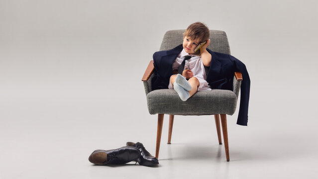 Little boy wearing oversized business suit sitting in armchair talking on smartphone. Concept of communication, future leadership, business ambitions and playful childhood imagination.