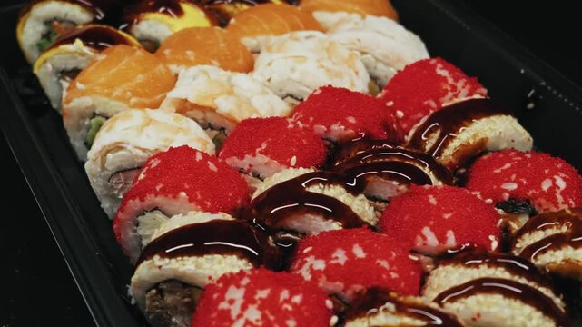 Variety of sushi rolls with salmon eel and red roe sit arranged in a black plastic container. Camera pans closely over the fresh Japanese cuisine highlighting glossy unagi sauce.