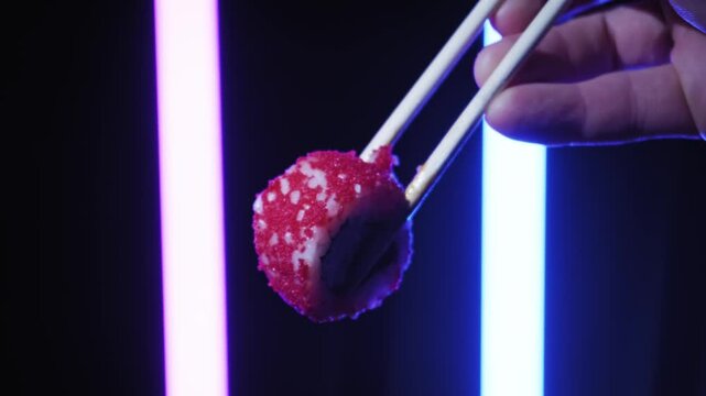 Male hand holds a California sushi roll with wooden chopsticks against dark backdrop. Vertical purple and blue neon tubes provide dramatic colorful cyber illumination.