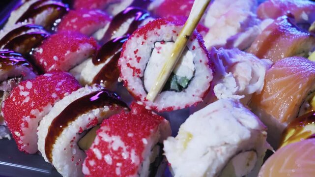 Wooden chopsticks lift sushi roll covered in red tobiko and cream cheese from platter. Bright purple and blue neon lights create vibrant nightclub atmosphere.