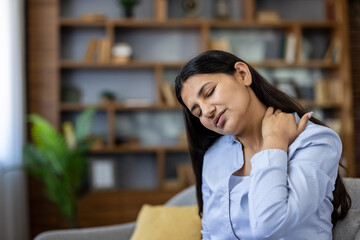 Young woman grimacing indoors, rubbing sore neck and shoulder from fatigue and poor posture,...