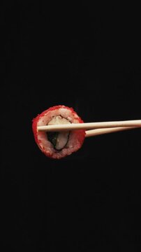 Male hand holds fresh California sushi roll coated in red tobiko using wooden chopsticks. Studio lighting highlights texture of rice and caviar against deep black background.