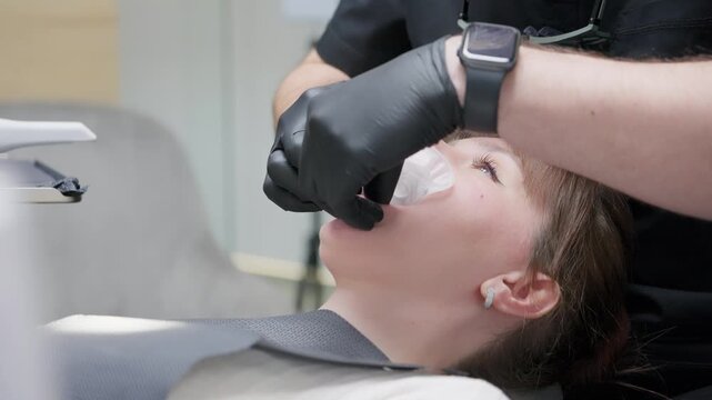 Professional orthodontist in a modern dental clinic wearing black gloves placing a cheek retractor in a young female patient's mouth to prepare for orthodontic examination or dental treatment