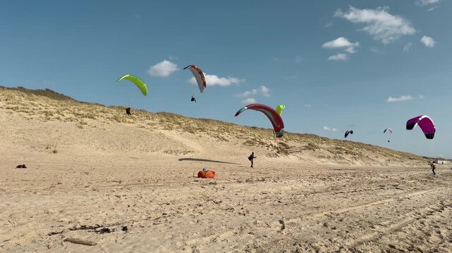 Action shot of multiple paragliders with colorful parachute wings soaring above sandy beach with dune landscape, participants on shore and spectators visible.