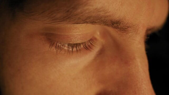 Male eye with long eyelashes blinks slowly in extreme close-up profile view. Warm golden lighting highlights skin texture and green iris detail against dark background.