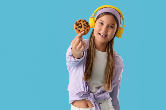 Cute teenage girl in headphones with delicious cookie on blue background