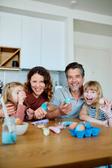 Happy family looking at camera while decorating Easter eggs at a modern kitchen table, celebrating...