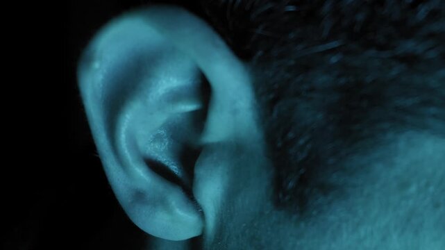 A macro close-up captures the details of a human ear illuminated by cool blue neon light against a black background. Concept of active listening, surveillance, and sensory perception.
