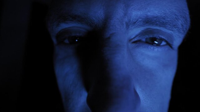 Male eyes stare intently at a screen reflecting blue light in total darkness. Cold illumination highlights the texture of the skin and creates a suspenseful mood.