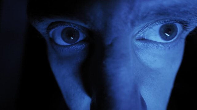 Distorted male face looks around frantically with wide darting eyes in extreme close-up. Eerie blue lighting creates a sinister atmosphere of paranoia and fear.