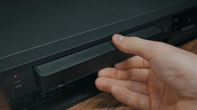 Male hand presses eject button and pulls black VHS cassette from VCR player. Retro technology sits on wooden surface with soft indoor lighting.