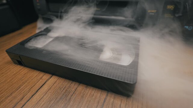 A black VHS cassette lies on a wooden surface as thick white smoke pours over it, obscuring the plastic casing. Concept of obsolete technology, data destruction, and nostalgic analog video.