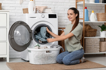 Beautiful young happy woman with basket taking clean laundry from washing machine at home