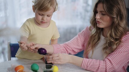 Naklejka premium Mother and child share joyful Easter egg activity. Child holds purple egg while mother assists with care. Soft sunlight bathes the cozy, warm indoor setting