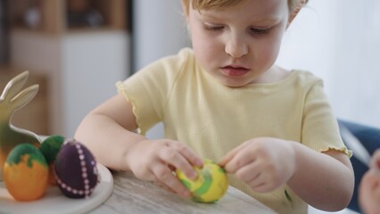 Fototapeta premium Little child focuses on colorful Easter eggs. Soft light highlights gentle concentration. Wooden table holds playful, decorated eggs. Child wears light yellow shirt, curls at collar