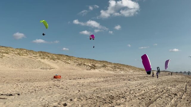Sky view of multiple paragliders with colorful parachute wings soaring high above sandy beach and dune landscape, participants visible on shore.