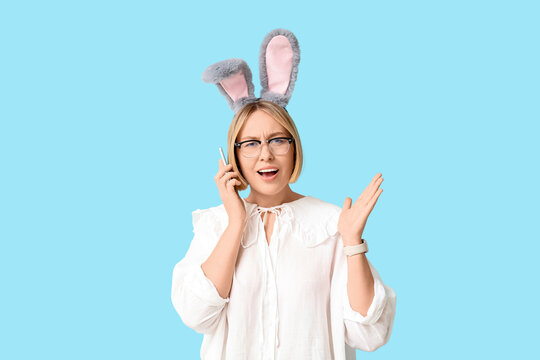 Worried adult businesswoman in bunny ears talking by mobile phone on blue background. Easter celebration