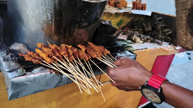 Street food vendor grilling chicken satay on traditional charcoal grill with smoke
