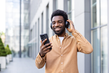 Young man smiling on an urban street, enjoying music and podcasts on wireless headphones while...