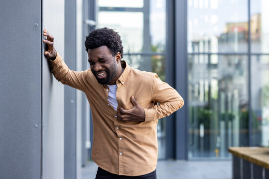 Adult individual experiencing sharp pains in their chest area, grasping their shirt while leaning against a wall outdoors, indicating discomfort and potential health suffering
