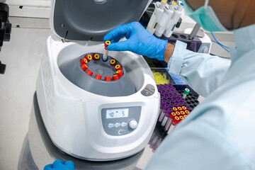 A lab technician wearing blue gloves carefully places a blood sample test tube into a clinical centrifuge machine for medical analysis in a laboratory setting © Leo Lintang