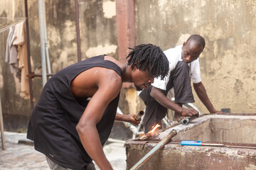 Two men working together to heat and repair a metal pipe near a concrete structure. Outdoor...