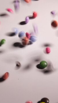 Vertical 4K 5x slow motion of colorful mini candy-coated chocolates falling and filling the frame against a white background. Cinematic lighting on a rainbow of sweets covering the screen.