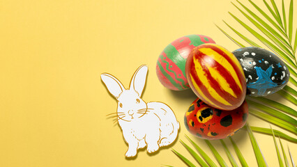 A festive arrangement featuring four hand-painted Easter eggs, a white bunny drawing, and a green palm leaf on a solid yellow background with ample copy space for text
