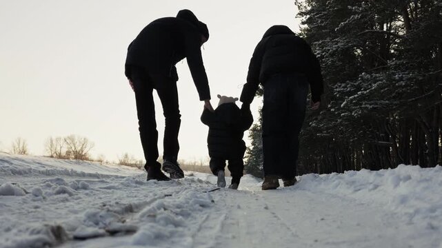 Parents and toddler walk away holding hands along a snowy road in a winter forest. Low angle camera follows boots stepping on frozen white ground.