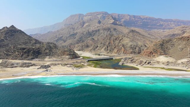 Aerial perspective of turquoise ocean water meeting a sandy coastline with a road bridge and rugged mountains in Salalah, Oman.