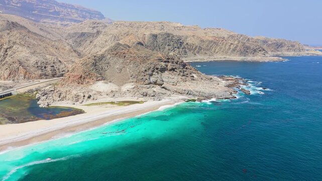 Aerial view of turquoise ocean waves crashing against a sandy beach bordered by mountains and a coastal highway in Salalah, Oman.
