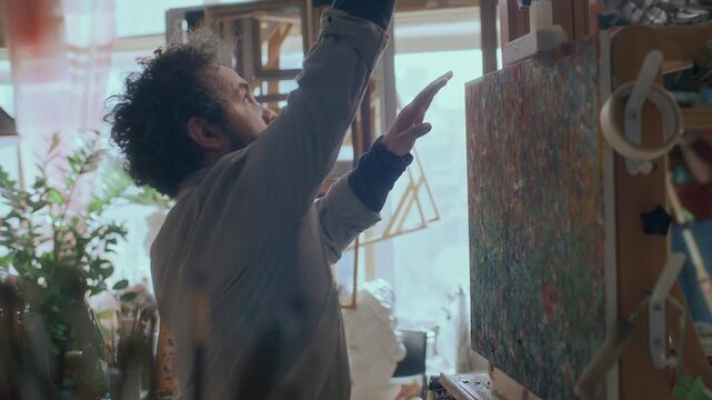 Male painter in stained apron walking into creative studio filled with artistic tools and setting up canvas on easel before starting work