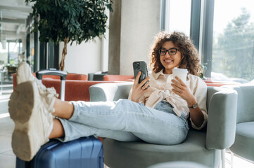 Relaxing on chair and using phone. Beautiful woman is in airport hall with suitcase