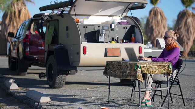 Mature woman sits by her teardrop camper trailer and works on her computer by the beach at Salton Sea in California.