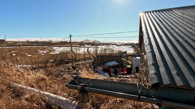 Rural farmhouse with farming equipment, dry grass, laundry and open landscape. Snow patches and clear blue sky create a natural countryside setting showing agricultural work and daily rural life.