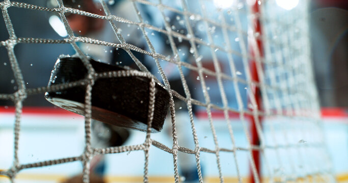 Close-up action shot of an ice hockey puck hitting the goal net during a scoring moment. Dynamic sports scene with ice spray and a blurred hockey player in the background.