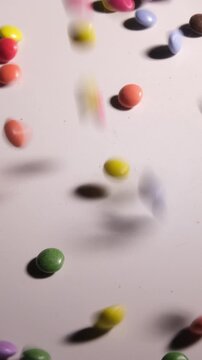Vertical 4K 5x slow motion of colorful mini candy-coated chocolates falling and filling the frame against a white background. Cinematic lighting on a rainbow of sweets covering the screen.