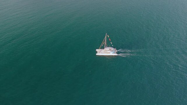 A white catamaran yacht cruises across calm blue ocean water, creating a noticeable wake. The high aerial view shows its clear path during daytime