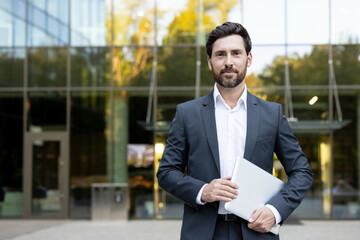 Naklejka premium Confident businessman standing outdoors holding a laptop, representing modern work, technology, success, and corporate professionalism in front of a glass office building