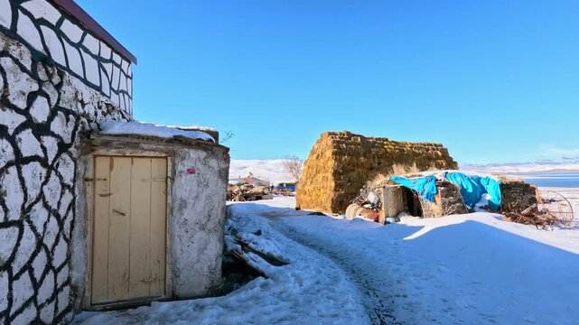 Snowy path in rural area leading between old structures and haystack toward a lake. Clear blue sky and winter light create a peaceful countryside scene with depth and natural composition.