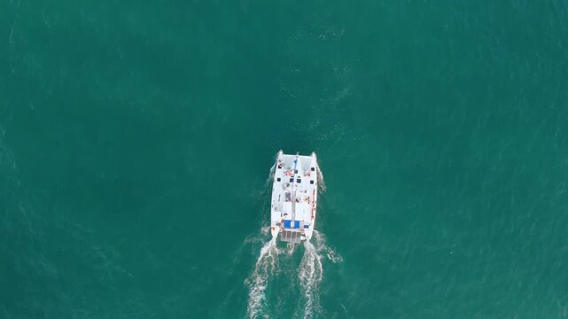 An aerial perspective shows a white catamaran boat cruising across clear teal ocean water. The boat leaves a noticeable foamy wake behind it