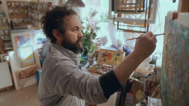 Male artist holding palette, mixing vibrant colors with brush and painting on canvas in sunlit creative studio filled with artworks, tools and frames
