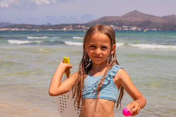 Young girl with long hair holding a beach toy and a colorful ball stands on the shore of a sunny beach with clear blue water and mountains in the background © Irina Flamingo