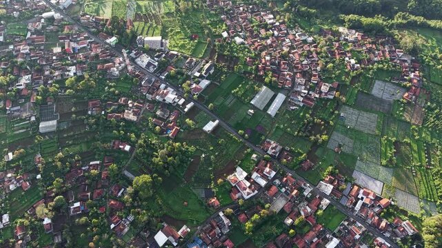 Sunlit aerial view of a rural village surrounded by patchwork farmland, green fields, roads and clustered rooftops, conveying peaceful countryside life, agricultural patterns and natural textures.