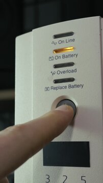 A male finger presses the power button on a white uninterruptible power supply unit to turn it on. Concept of electrical safety, backup energy solutions, and data protection technology.