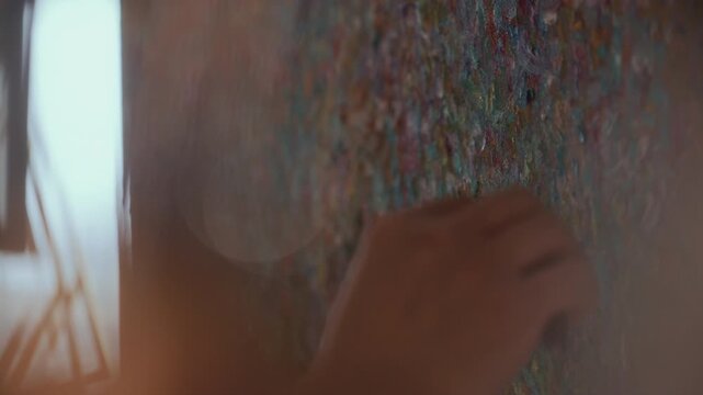 Artists hand using palette knife to apply thick vibrant oil paint to heavily textured canvas, working in impasto technique on contemporary abstract artwork in sunlit studio. Close up view