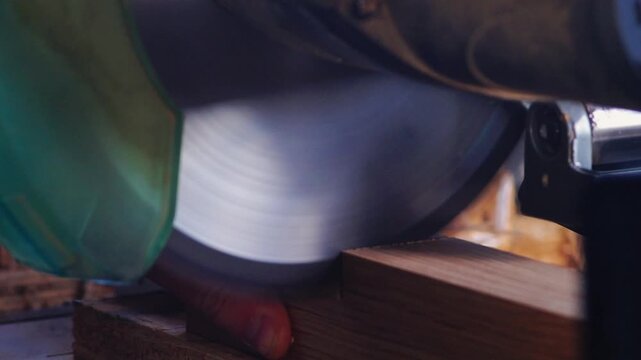 Close up of carpenter hands holding oak wood while a mitre saw blade spins and cuts through the hardwood in a workshop. Carpentry woodworking process and DIY home improvement concept.